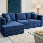 Luksusowy salon z niebieską, pikowaną sofą M125 z weluru, ustawioną przy szklanym stoliku kawowym, z dekoracyjnym panelem i nowoczesną lampą w tle.