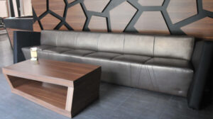 Sofa M025 w ciemnoszarej tapicerce stojąca przed ścianą z czarnymi geometrycznymi panelami i drewnianym tłem, z drewnianym stolikiem kawowym na pierwszym planie.