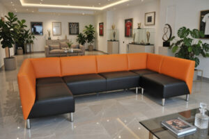 Narożnik M027 w pomarańczowo‑czarnych kolorach stojący w nowoczesnym lounge z marmurową podłogą, trofeami w gablotach i sportowymi koszulkami na ścianie.