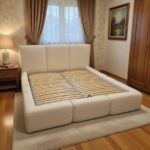 Łóżko L609 z białym, gładkim wezgłowiem i tapicerowaną ramą, pokazane bez materaca, z widocznymi drewnianymi listwami w eleganckiej sypialni.