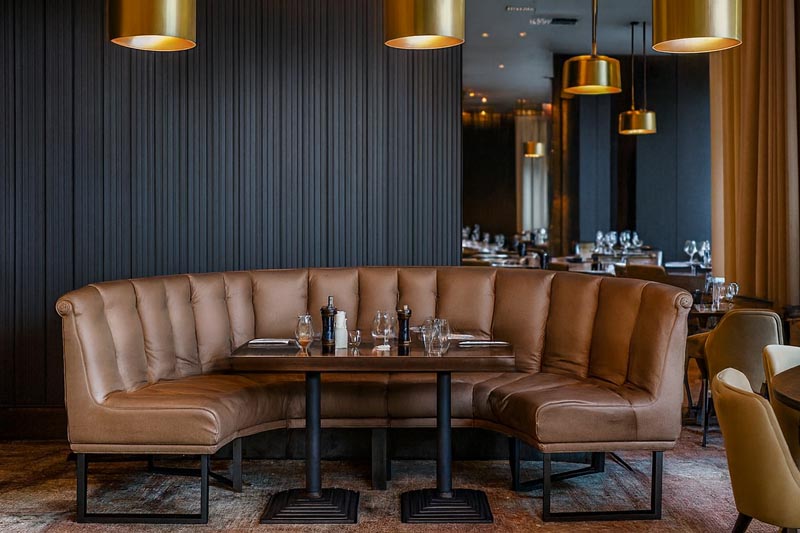 Loża M183 w brązowej tapicerce, otaczająca dwa stoły w nowoczesnej restauracji z ciemnymi panelami, lustrami i złotymi lampami nad strefą siedzącą.
