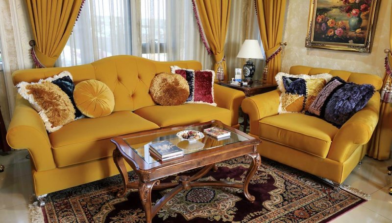 Elegancki salon z zestawem wypoczynkowym M441 od Mebel Akimo w musztardowym odcieniu, z dekoracyjnymi poduszkami i drewnianym stolikiem na tle dużych okien z zasłonami.