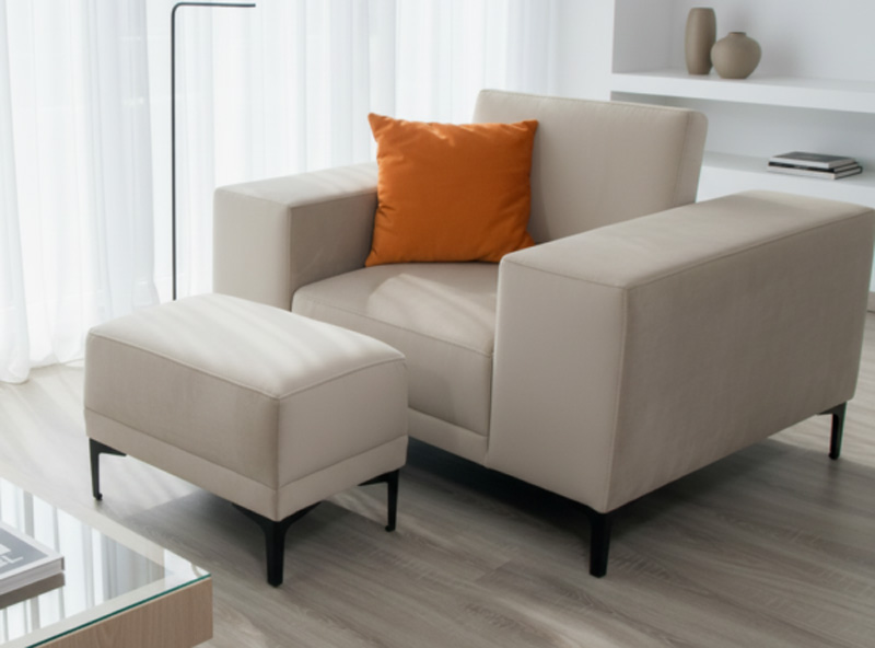 Fotel M006 firmy Mebel Akimo w beżowej tapicerce z podnóżkiem i pomarańczową poduszką, ustawiony w jasnym, minimalistycznym salonie.